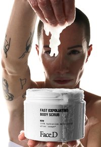 White container of fast exfoliating body scrub with textured lid, held above a person's hand, displaying a creamy scrub. Black text on front.