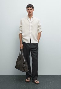 Herrliches Leinenhemd, hellbeige, mit langen Ärmeln und Knopfverschluss. Kombiniert mit dunkelgrauen Hosen. Hält eine dunkelbraune Wildledertasche; trägt schwarze Flip-Flops.