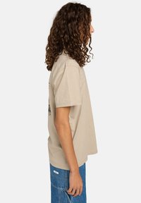 Camiseta de algodón color beige con mangas cortas, de corte relajado, y una vista lateral que muestra un diseño gráfico en la espalda. Combinada con pantalones de mezclilla azules.