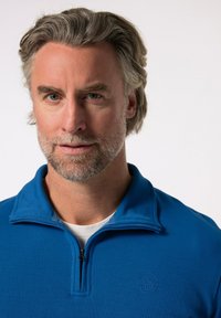 Modrý zipový svetr s vroubkovanou texturou a stojáčkovým límcem; má vyšívané logo na hrudi a viditelný bílý tričko pod ním.