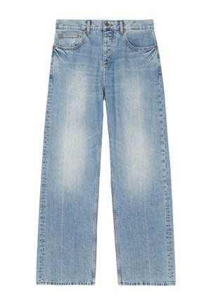 Homme portant une chemise noire à manches longues, un jean bleu clair, des chaussures noires et un bracelet argenté, debout devant un fond blanc uni.