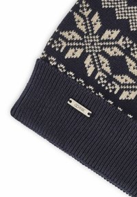 Granatowa zimowa czapka dziana z kremowymi wzorami śnieżynek, prążkowany mankiet oraz mała metalowa metka z logo przymocowana z boku.