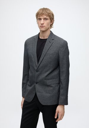 Junger Mann mit kurzen blonden Haaren, der einen grau karierten Blazer über einem schwarzen Hemd und schwarzen Hosen trägt, steht vor einem schlichten weißen Hintergrund.