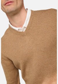 Beiger V-Ausschnitt-Pullover aus strukturiertem Stoff. Mit geripptem Kragen und subtilen Nähdetails. Über einem weißen Hemd getragen.