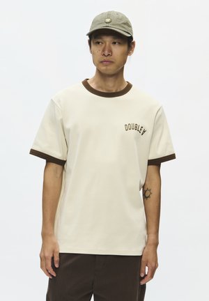 Uomo che indossa una maglietta color crema con bordino marrone e testo "DOUBLE A", un cappellino verde, pantaloni scuri e un piccolo tatuaggio del sole sull'avambraccio sinistro.