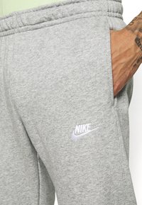 Sivé teplákové nohavice vyrobené z mäkkej tkaniny, s elastickým pásom a bočnou vreckom, na nohe je vyšitý biely logo Nike.