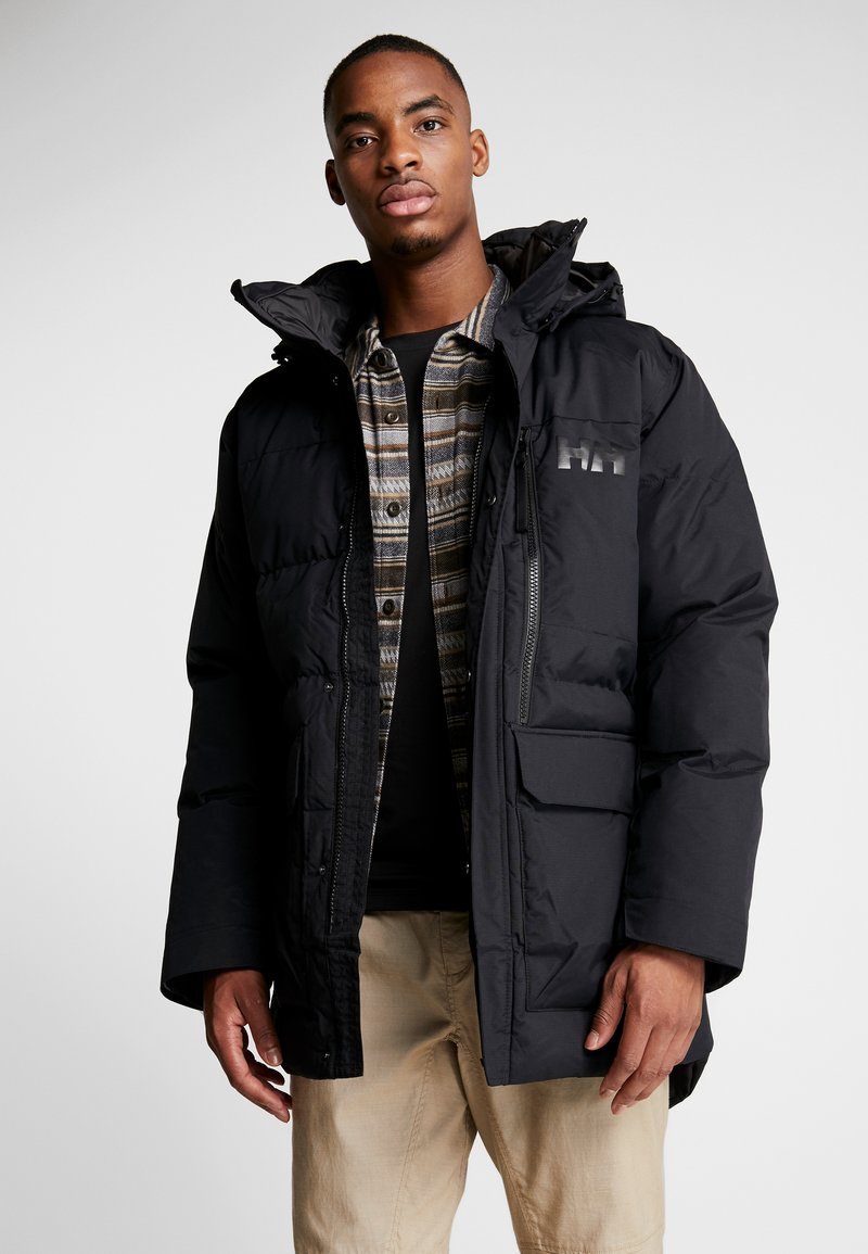 Black puffer jacket with a hood, featuring a front zip and pockets. Paired with a plaid button-up shirt and beige pants.