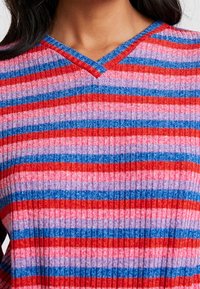 Prążkowany sweter w odcieniach różu, czerwieni i niebieskiego. Posiada dekolt w kształcie litery V oraz prążkowaną fakturę z naprzemiennymi szerokimi i wąskimi paskami.