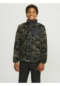 Fleece bunda v černé barvě s krémovým paisley vzorem, zahrnující kapsu na zip na hrudi a vysoký límec, v kombinaci s tmavými kalhotami.