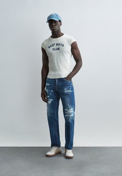 Homme portant un débardeur blanc "Bad Boys Club", un jean bleu déchiré, des baskets blanches et une casquette de baseball en denim bleu clair, debout contre un mur uni.