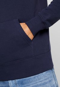 Felpa blu navy con finitura testurizzata, dotata di tasca frontale e polsini a coste. Abbinata a jeans blu chiaro, evidenziando un design casual.