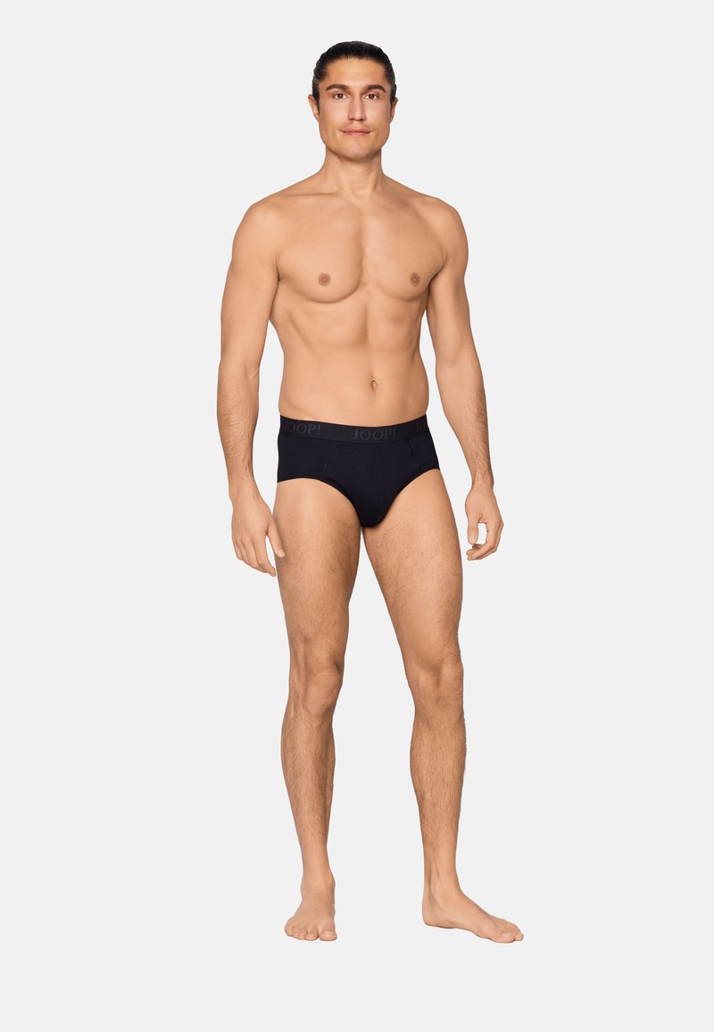 Homme debout pieds nus devant un fond uni, portant un slip noir avec un logo sur la ceinture, face à l'avant avec une posture détendue.