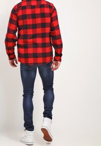 Camisa de franela a cuadros roja y negra, vaqueros ajustados azules y zapatillas blancas. La camisa tiene cuello abotonado y mangas largas.