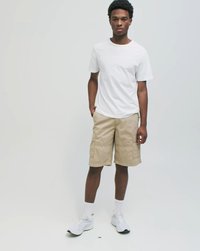 Joven de pie vistiendo una camiseta blanca sencilla, pantalones cortos cargo beige, calcetines blancos y zapatillas blancas contra un fondo gris claro.