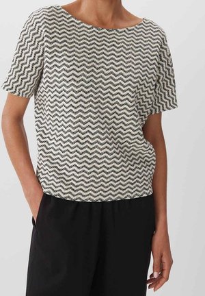 Femme portant un haut blanc à manches courtes avec un motif en zigzag ondulé noir et un pantalon noir, une main dans la poche, sur un fond uni.