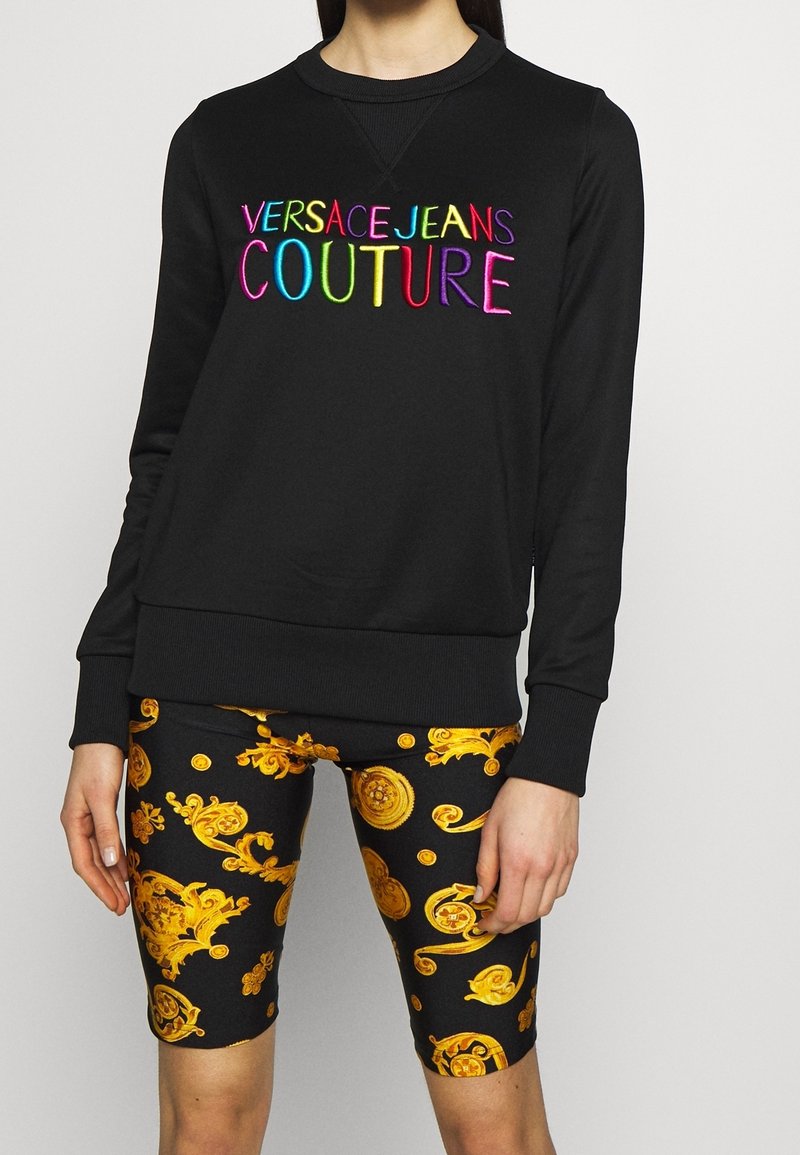 Sweat-shirt noir avec texte coloré « Versace Jeans Couture » associé à un short noir et or à motif baroque longueur genoux.