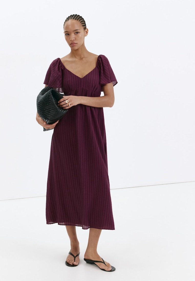 Femme aux cheveux tressés portant une robe midi violette à manches bouffantes, tenant une pochette noire texturée et portant des tongs noires, debout sur fond blanc.