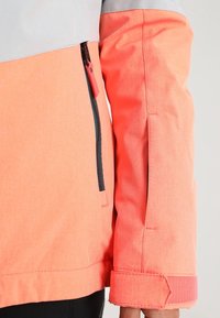 Chaqueta resistente al agua con un llamativo esquema de color coral y gris. Presenta un bolsillo con cremallera y mangas ajustables. Textura suave.