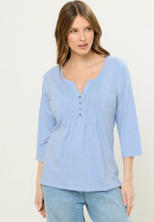 Kobieta ubrana w jasnoniebieską bluzkę z rękawem 3/4, zdobioną marszczeniami i guzikami z przodu, w połączeniu z jasnoniebieskimi dżinsami, na białym tle.
