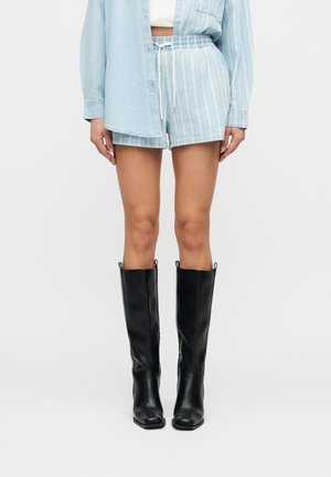 Mujer con pantalones cortos de rayas azul claro y camisa a juego, con botas de cuero negras hasta la rodilla, de pie frente a un fondo blanco.