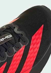 Black athletic shoe with red geometric patterns, textured fabric upper, thick black sole, and black laces. Features logo on the tongue.