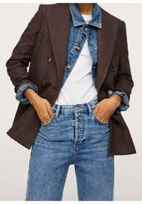 Bruine houndstooth dubbelbreasted blazer gedragen over een blauwe denim jas, gecombineerd met lichtgekleurde denim jeans met hoge taille. Knopen zichtbaar.