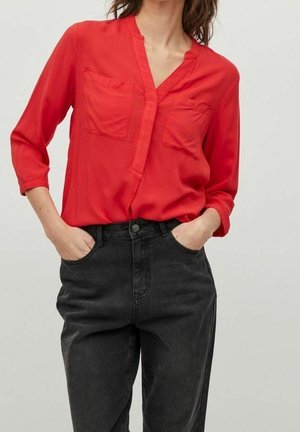 Kvinde iført en knaldrød langærmet bluse med brystlommer og højtaljede sorte denimjeans, hænderne afslappet i lommerne.