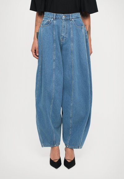 Weite blaue Denim-Jeans mit vertikalen Nähten, hoher Taille, kombiniert mit schwarzen spitzen Schuhen und tätowierten Armen, die vor einem weißen Hintergrund sichtbar sind.