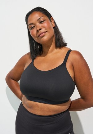 Frau mit mittel-dunkler Haut und schulterlangem schwarzen Haar, die einen schwarzen Sport-BH und hochgeschnittene Leggings trägt, steht mit den Händen am Rücken.