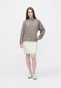 Brun strikket sweater med rund hals kombineret med hvid lædernederdel. Modellen står med den ene hånd på hoften og har lette slip-on sko på.