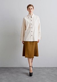 Biela košeľa s golierom, skombinovaná s hnedou sukňou po kolená. Modelka má na sebe čierne platformové topánky a stojí na jednoduchom pozadí.