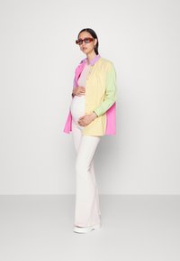 Camisa multicolorida em rosa, amarelo e verde, usada sobre um top justo bege; calças brancas largas e óculos de sol vermelhos. Texturas suaves, design casual.