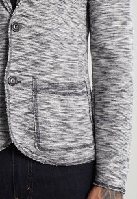Blazer de punto texturizado en gris y blanco con dos botones, un bolsillo en el pecho y corte entallado, que presenta un dobladillo ligeramente deshilachado.