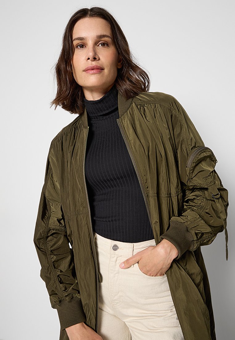 Kobieta z brązowymi włosami do ramion nosi oliwkową zieloną kurtkę, czarny prążkowany golf i beżowe spodnie, stojąc na jasnym tle.