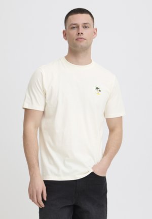 Uomo che indossa una T-shirt a maniche corte color avorio con una piccola grafica di palma e isola sul petto sinistro, abbinata a pantaloni neri.