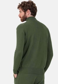 Homme aux cheveux bouclés et à la barbe, portant un pull à col roulé vert foncé et un pantalon assorti, les mains dans les poches, faisant face à gauche, fond gris.
