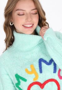 Mintgrüner Kabelstrickpullover mit hohem Kragen. Verziert mit bunten, flauschigen Buchstaben und einem Herzen auf der Vorderseite. Weiches, strukturiertes Material.