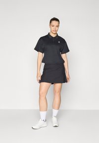 Juoda polo marškinėliai trumpomis rankovėmis, su mažuoju logotipu, derinami su juoda klostuota sijonu. Apsirengta su baltais sportiniais bateliais ir kojinėmis.