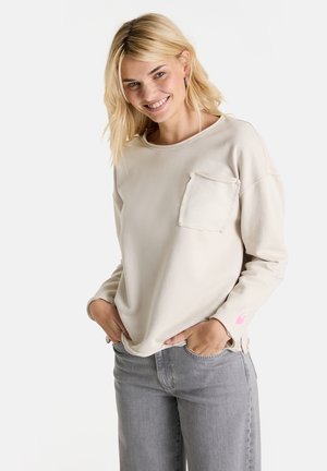 Blonde Frau lächelt, trägt ein cremefarbenes Langarmshirt mit einer Brusttasche und hellgraue Jeans, die Hände in den Taschen, vor einem weißen Hintergrund.