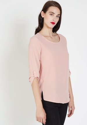 Mujer con cabello oscuro y labios rojos, vistiendo una blusa rosa clara con mangas de 3/4 remangadas y pantalones negros, de pie contra un fondo blanco.