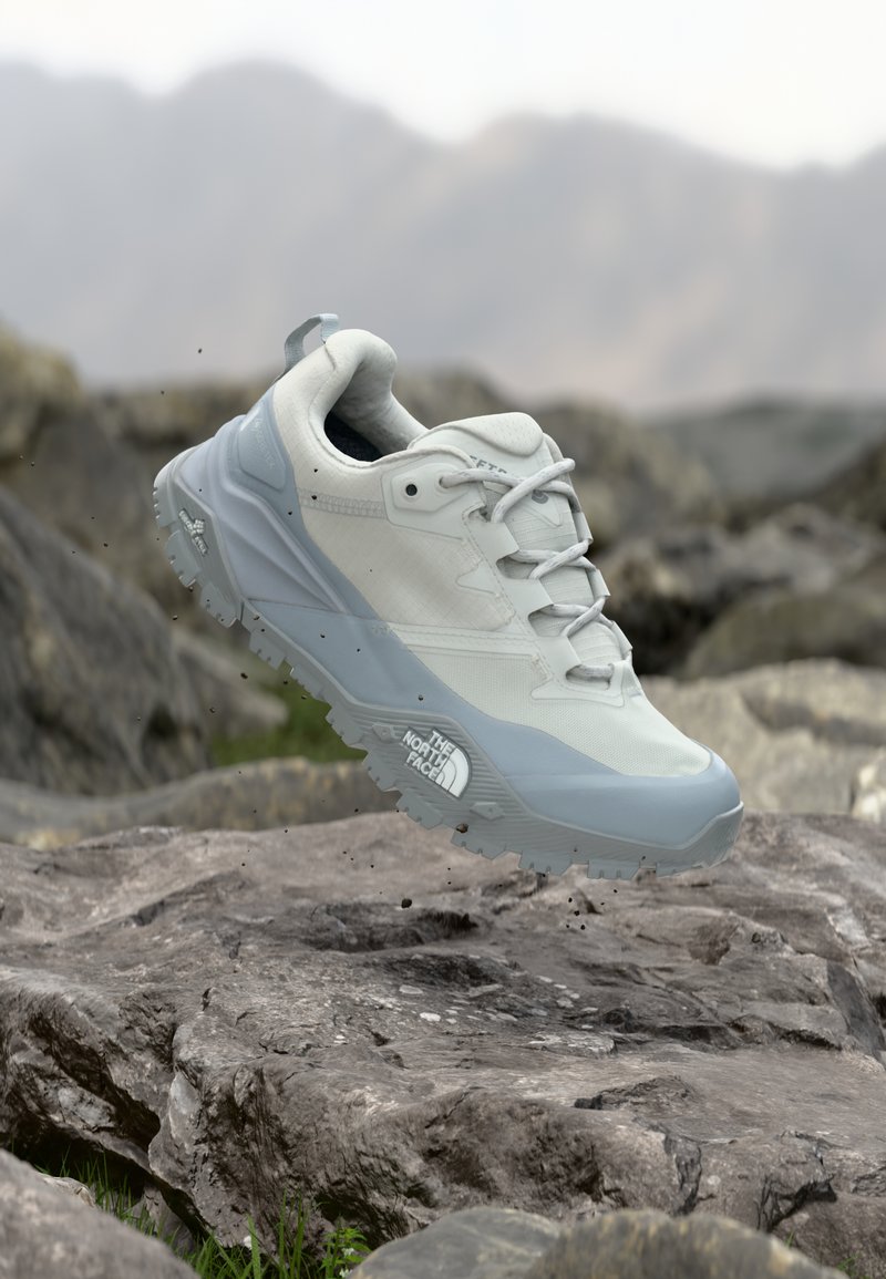 Άσπρο και γκρι πεζοπορικό παπούτσι The North Face αιωρείται πάνω από βραχώδες έδαφος με φόντο το βουνό και διασκορπισμένα σωματίδια χώματος.