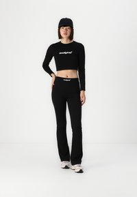 Top noir à manches longues et cropped, associé à un pantalon taille haute avec des accents de logo. Le modèle porte une casquette noire et des chaussures de sport roses. Texture plate.