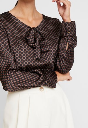 Femme portant un chemisier noir à manches longues avec un petit motif circulaire et un col à nœud papillon, associé à un pantalon blanc taille haute avec bouton.