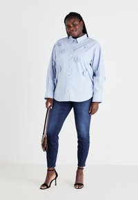 Egy világoskék csíkos inget viselő nő, melyen szövetvirágok találhatóak, sötét skinny farmert, fekete magassarkú szandált hord, és egy barna táskát tart a kezében.