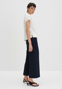 Haut blanc à manches courtes avec fentes latérales, associé à un pantalon large bleu marine et des sandales noires, sur un fond clair.