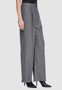 Donna che indossa pantaloni grigi a vita alta e gamba larga con pieghe frontali e stivali neri con tacco a punta, su sfondo neutro.