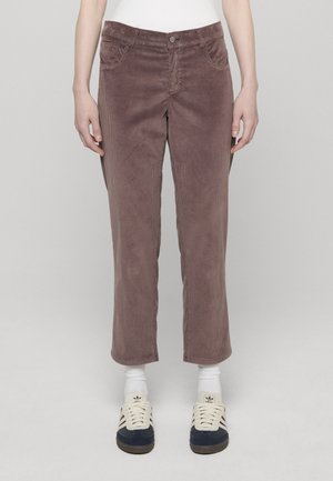 Henkilö, joka pukeutuu vaaleanvioletteihin samettihousuihin, valkoisiin sukkiin ja tummansinisiin ja valkoisiin Adidas-tennareihin, seisoo yksinkertaisen vaaleanharmaan taustan edessä.