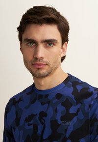 Giovane uomo con capelli corti castani e leggera barba incolta, che indossa una camicia a fantasia mimetica blu e nera, che guarda direttamente in avanti su uno sfondo semplice.