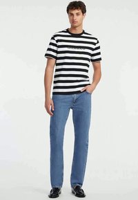 Jovem vestindo uma t-shirt preta e branca com listras horizontais, jeans azuis e sapatos pretos, de pé com uma mão no bolso, em fundo branco.