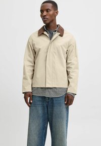 Beige arbejdsjakke med en brun krave, der har en frontlynlås og sidelommer, båret over en ternet skjorte og brede jeans.